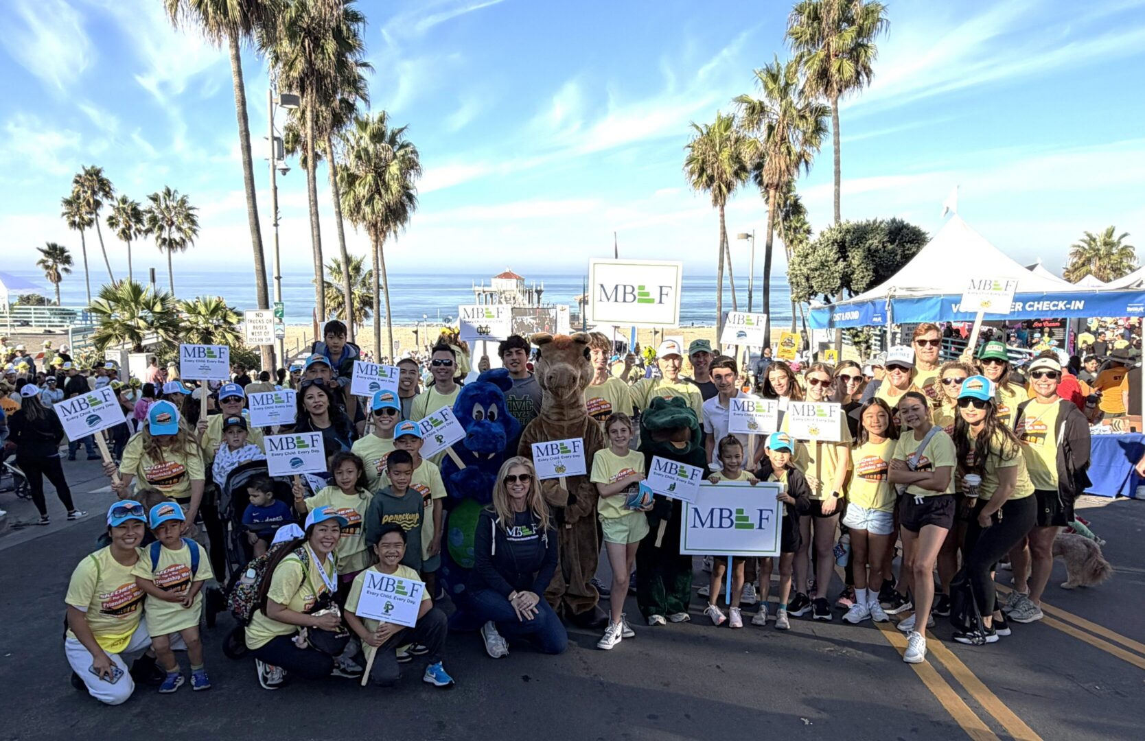A large group of people gathered outdoors near palm trees and the beach.